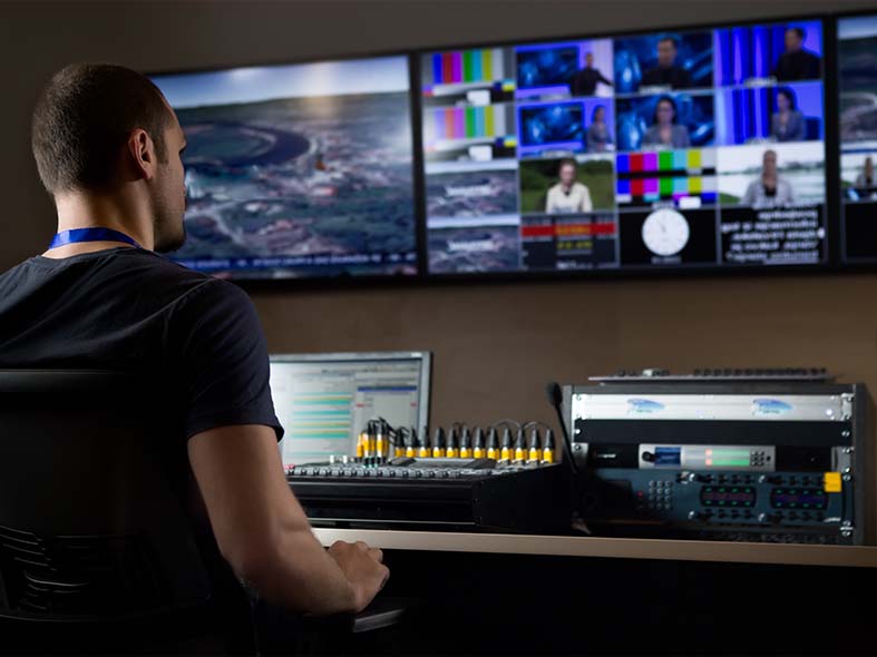 Man sits behind media equipment with a soundboard and split-screen TV, working as the chosen solutions provider for media companies.